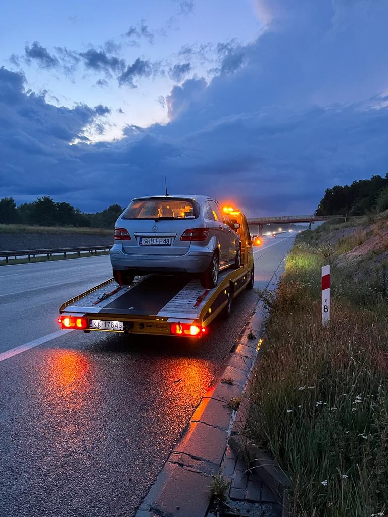Srebrny samochód osobowy z rejestracją SRB FP48 przewożony na żółtej lawecie z włączonymi światłami ostrzegawczymi na poboczu autostrady o zmierzchu.