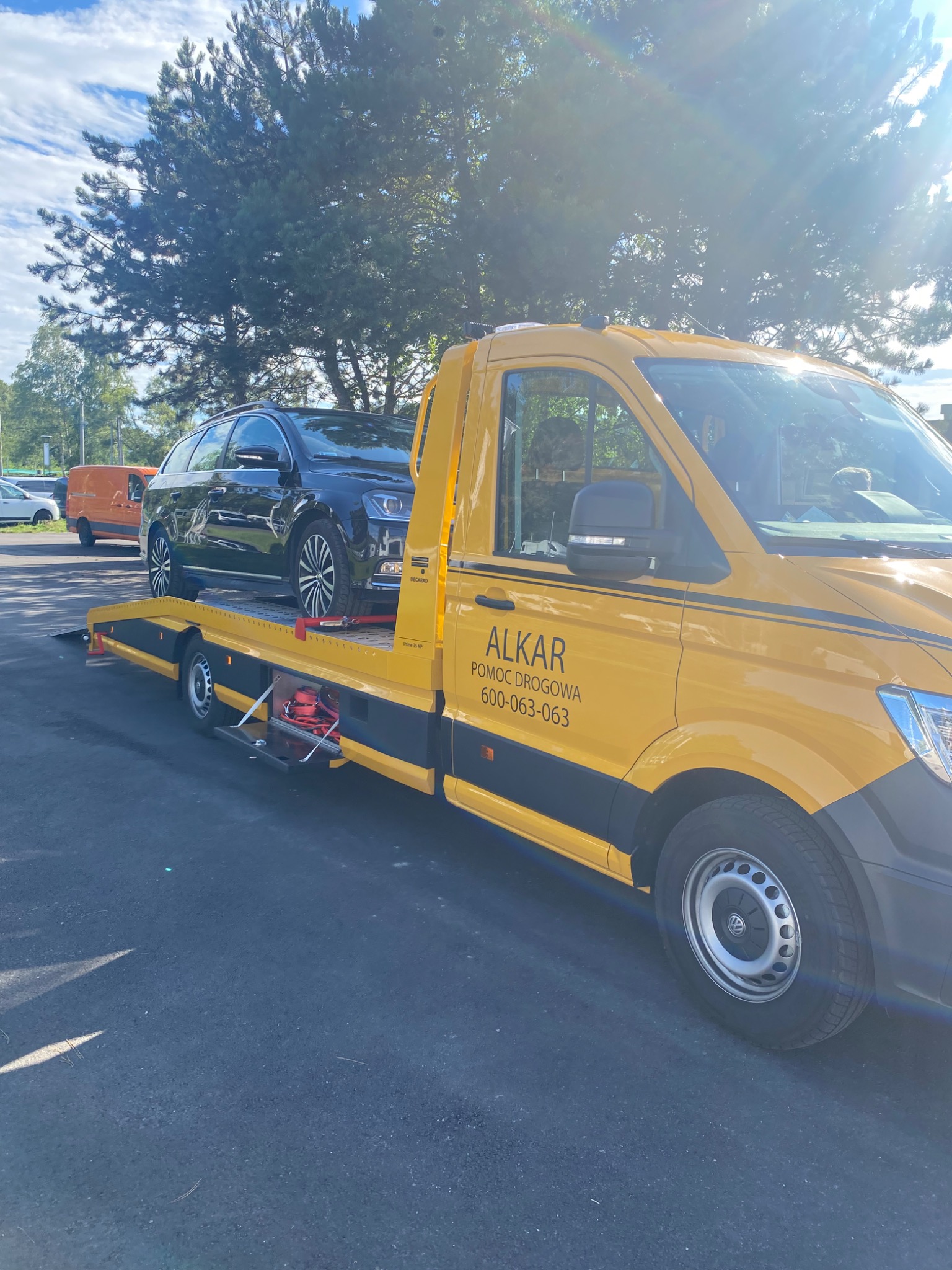 Żółta laweta z czarnym samochodem osobowym na platformie, widoczny napis 'ALKAR Pomoc Drogowa' i numer telefonu, zaparkowana na parkingu w słoneczny dzień.