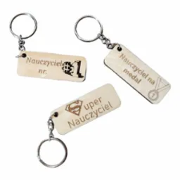 Trzy drewniane breloczki z grawerowanymi napisami 'Nauczyciel nr. 1' z pikselowym numerem, 'Super Nauczyciel' z symbolem Supermana oraz 'Nauczyciel na medal' z rysunkiem medalu.