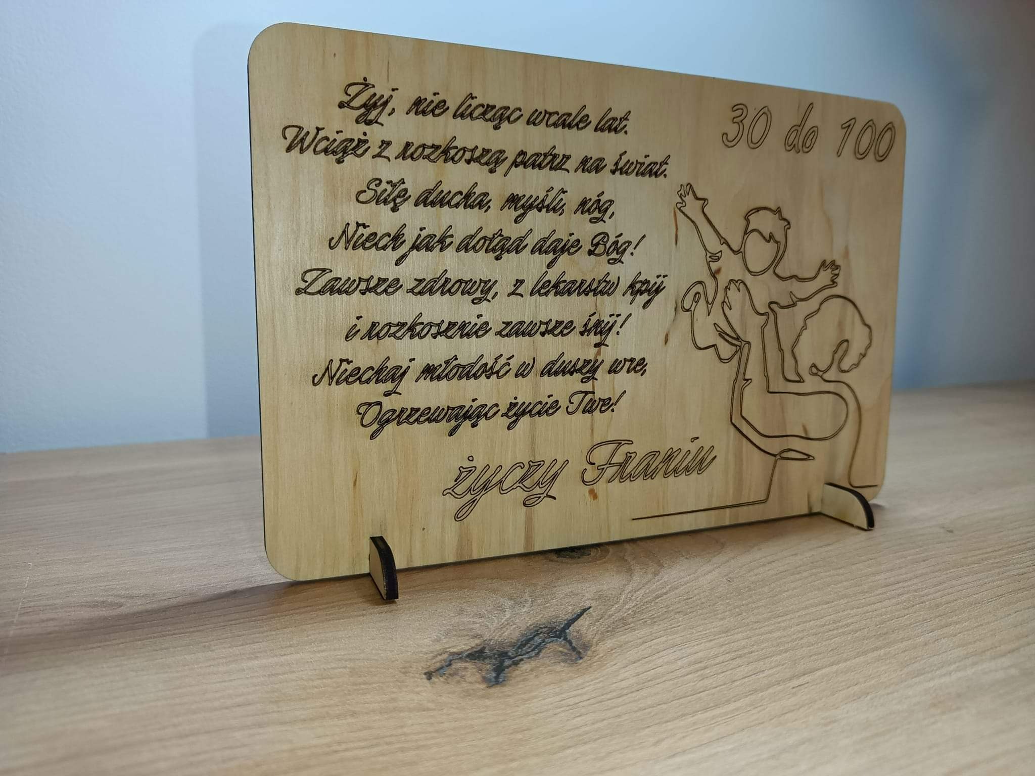 Drewniana tabliczka z wygrawerowanym laserowo tekstem życzeń urodzinowych i rysunkiem, stojąca na drewnianym blacie.