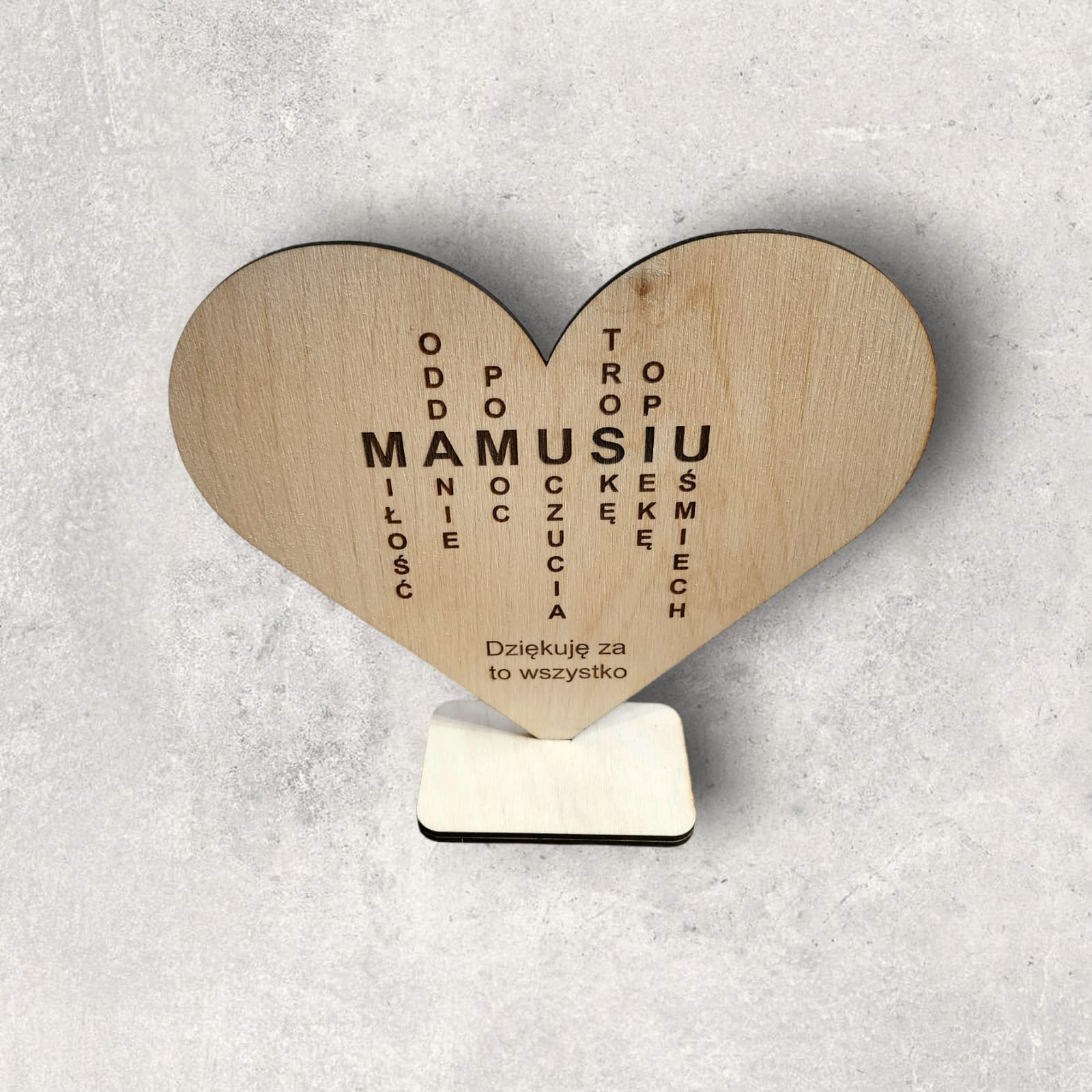 Drewniane serce z wygrawerowanym napisem 'Mamusiu' w formie krzyżówki słownej, umieszczone na drewnianej podstawce, na neutralnym szarym tle.