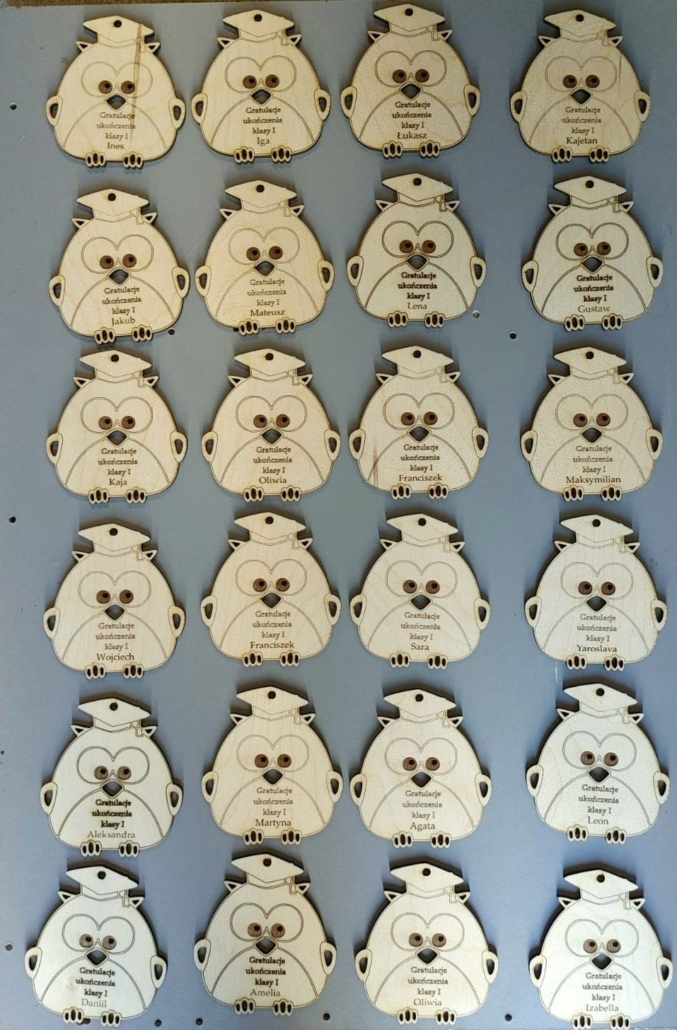 Drewniane zawieszki w kształcie sowy z wygrawerowanymi imionami i napisem 'Gratulacje ukończenia klasy I', ułożone w rzędach na szarym tle.