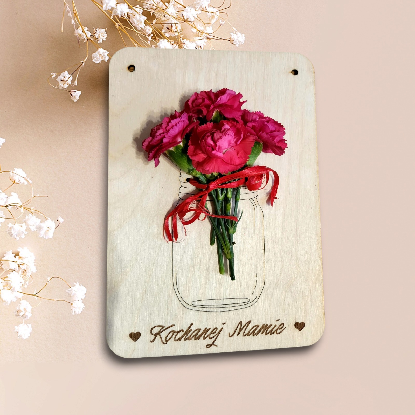 Drewniana kartka z wygrawerowanym słoikiem i napisem 'Kochanej Mamie', ozdobiona bukietem różowych goździków i czerwoną wstążką, na beżowym tle z gałązkami gipsówki.