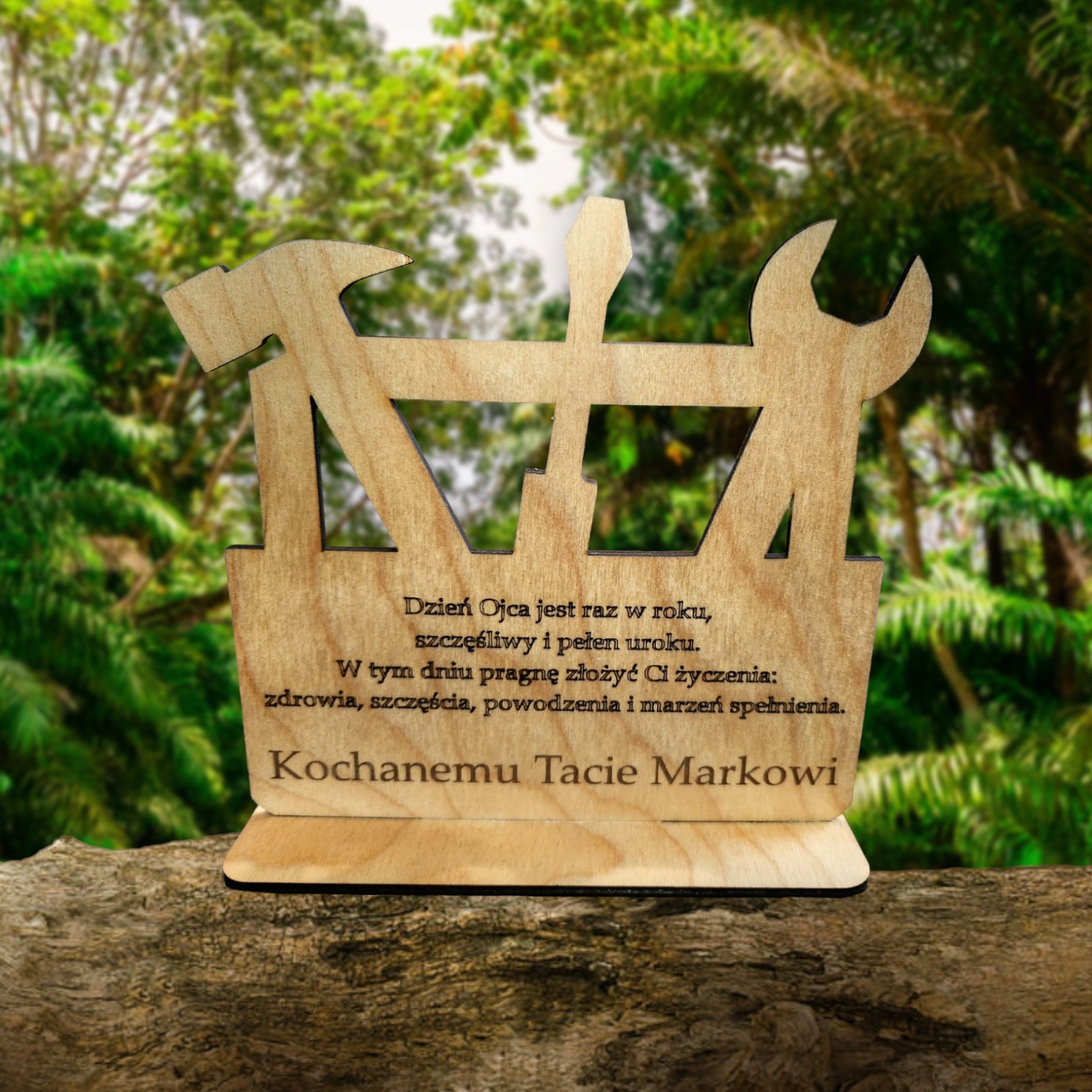 Drewniana statuetka z narzędziami (młotek, śrubokręt, klucz) i grawerowanymi życzeniami dla Taty Marka, umieszczona na pniu drzewa, na tle zieleni. Pamiątka z okazji Dnia Ojca od firmy z Koszalina.