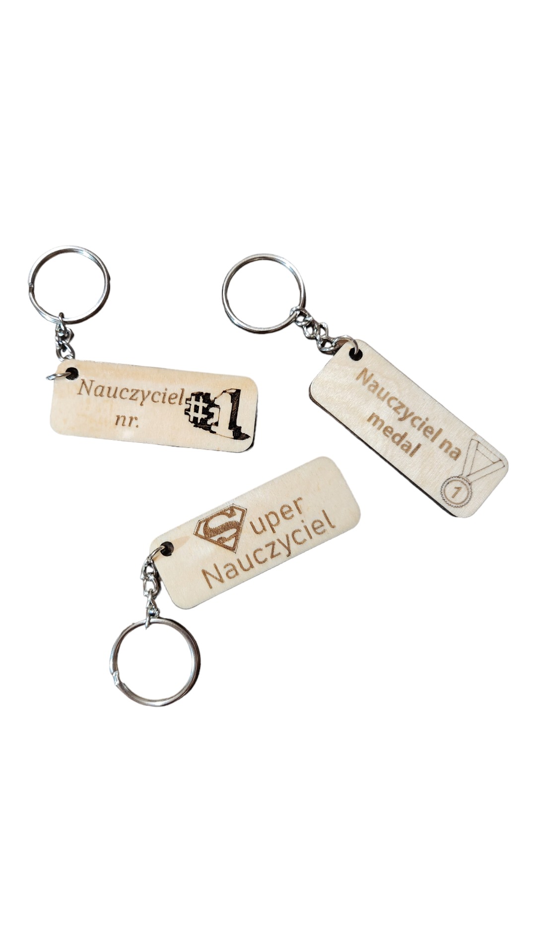 Trzy drewniane breloczki z grawerowanymi napisami 'Nauczyciel nr. 1' z pikselowym numerem, 'Super Nauczyciel' z symbolem Supermana oraz 'Nauczyciel na medal' z rysunkiem medalu.