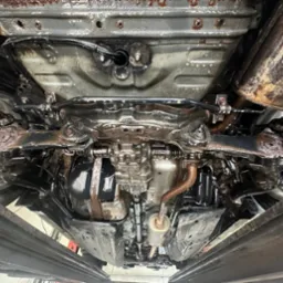 Podwozie samochodu po konserwacji antykorozyjnej, widoczne elementy zawieszenia, układu wydechowego i podłogi pokryte ochronną, błyszczącą powłoką. Ujęcie od dołu.