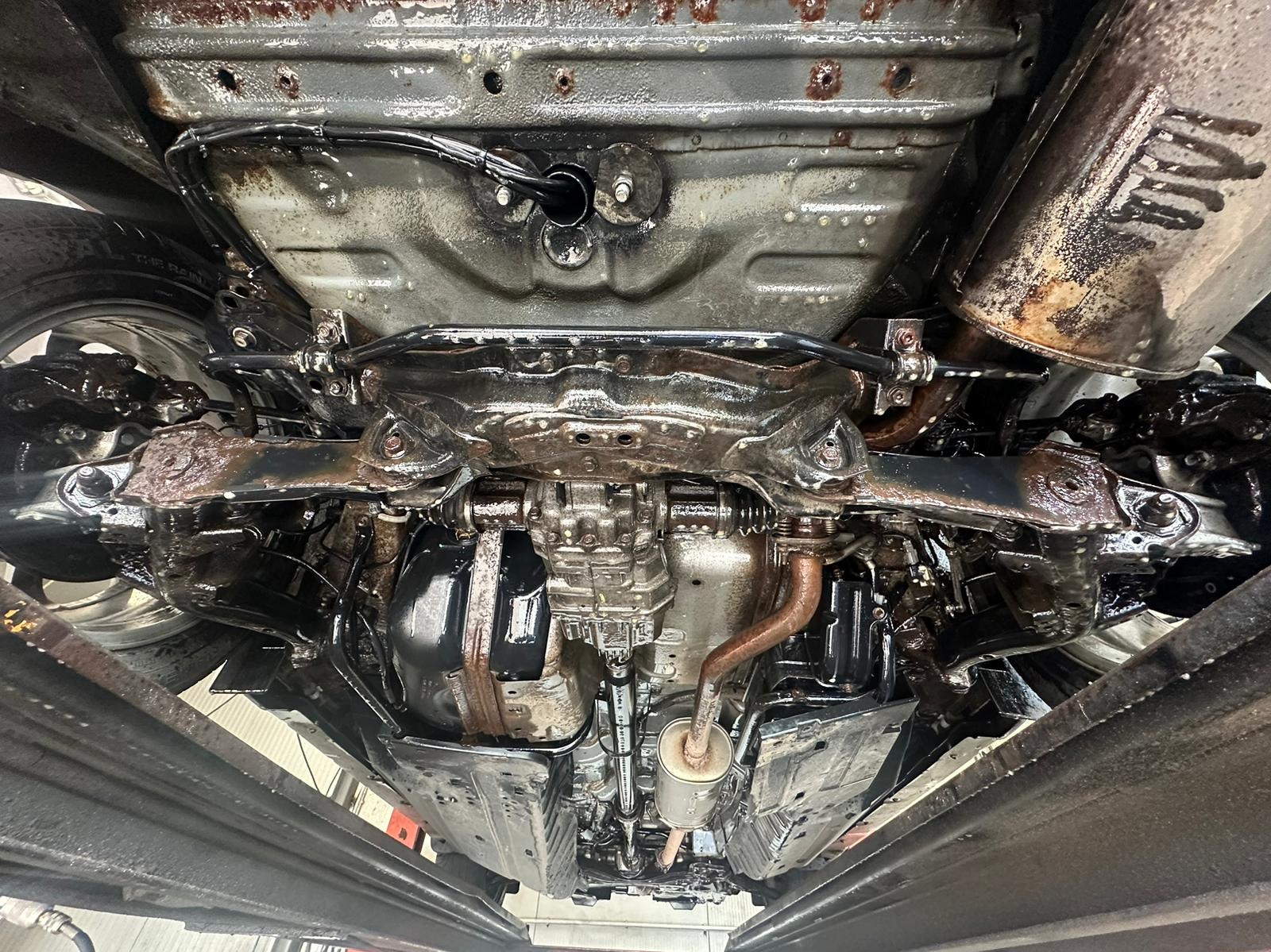 Podwozie samochodu po konserwacji antykorozyjnej, widoczne elementy zawieszenia, układu wydechowego i podłogi pokryte ochronną, błyszczącą powłoką. Ujęcie od dołu.