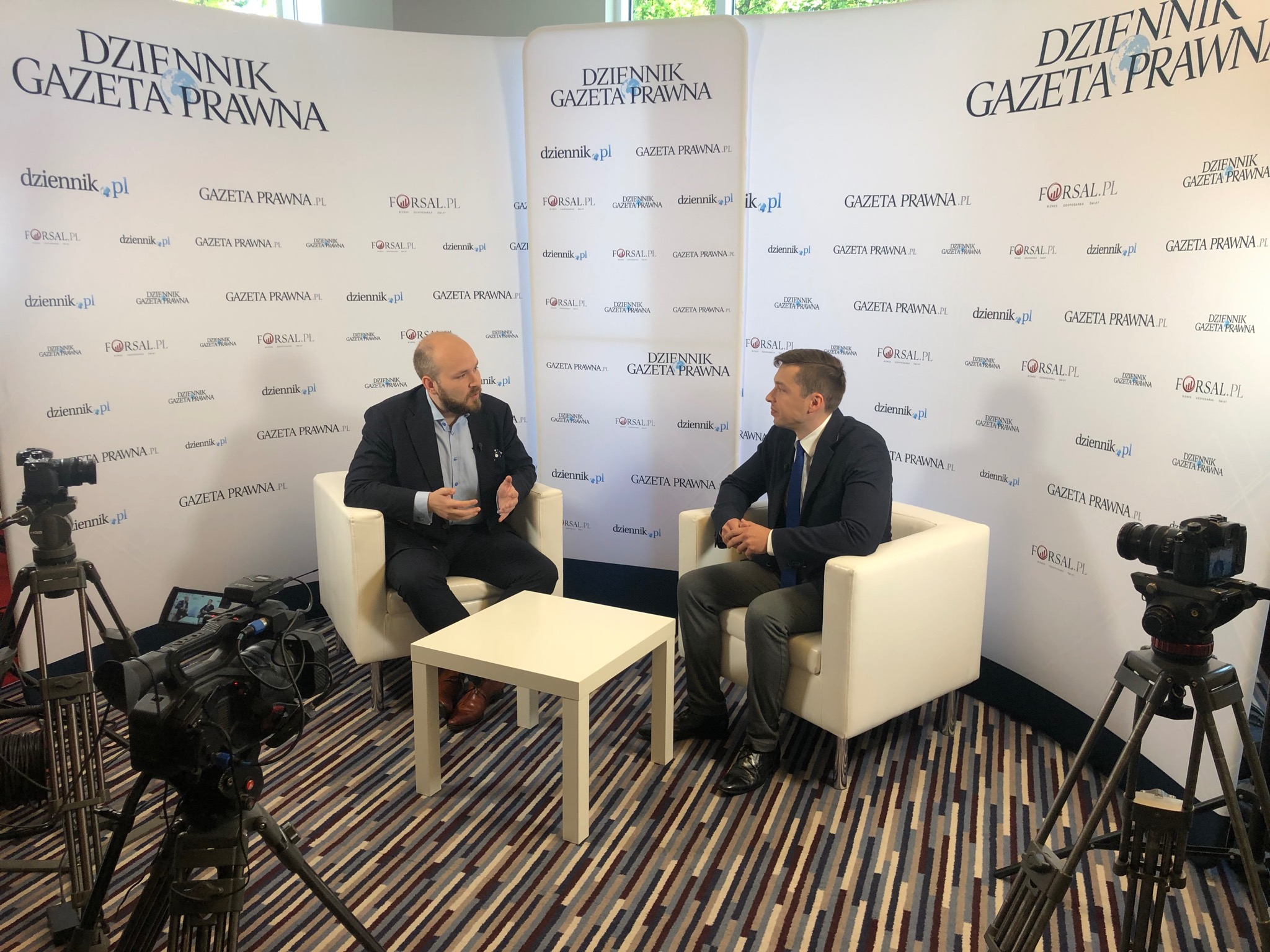 Radca prawny udziela wywiadu dla Dziennika Gazety Prawnej; dwóch mężczyzn siedzi w fotelach na tle ściany z logo dziennika, przed nimi stolik, widoczne kamery na statywach.