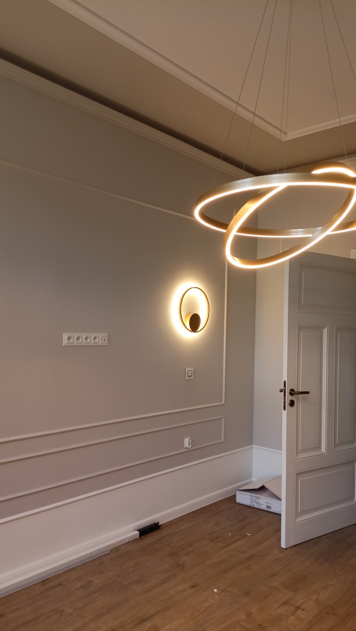 Nowoczesne oświetlenie: okrągły żyrandol i kinkiet z podświetleniem na szarej ścianie z dekoracyjnymi listwami, widoczne gniazdka elektryczne i drewniana podłoga, częściowo otwarte białe drzwi.
