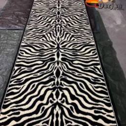 Długi dywan w czarno-biały wzór zebry, leżący na ciemnej, pofałdowanej folii ochronnej. W prawym górnym rogu widoczne logo firmy z karykaturalną postacią.