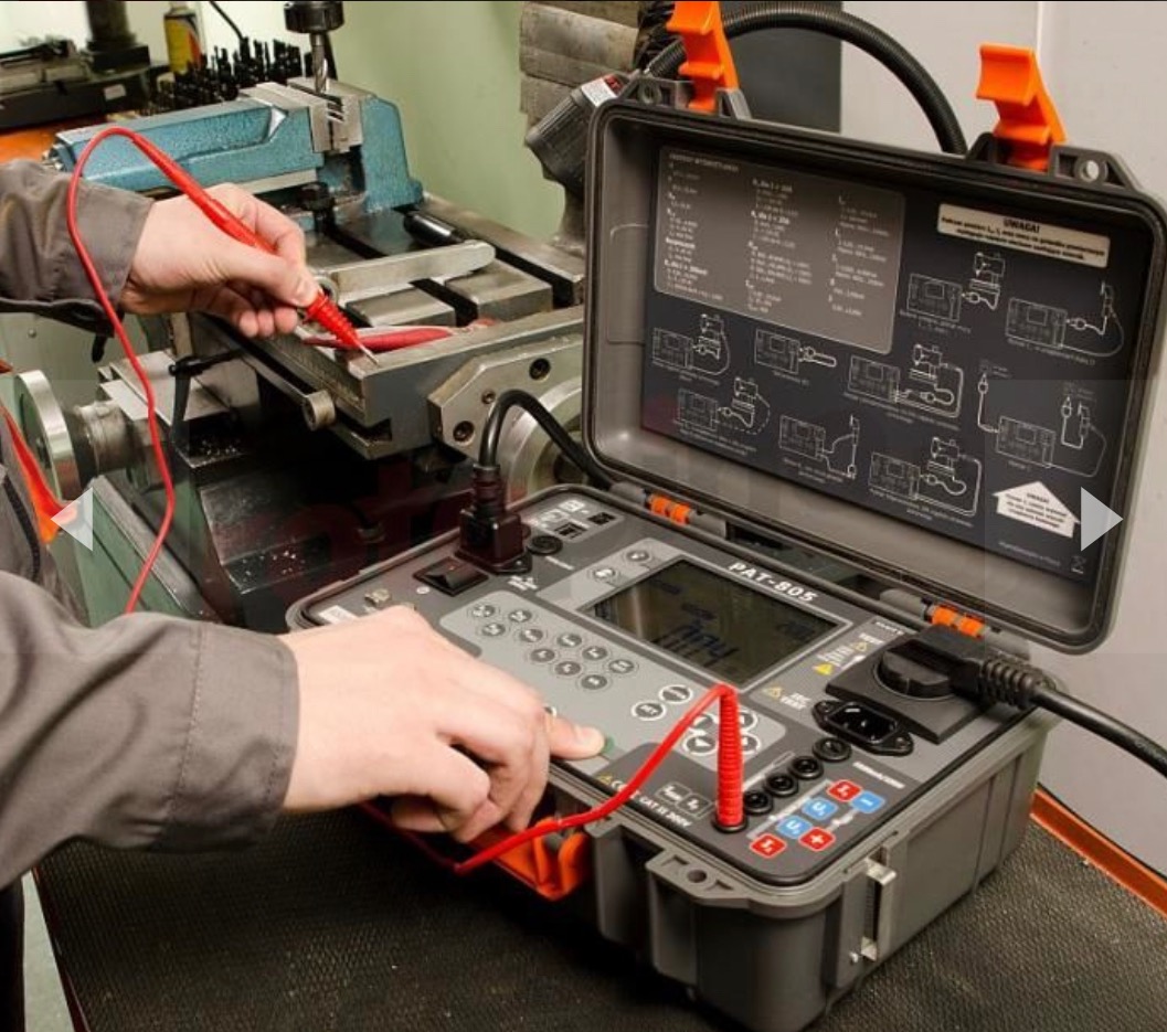 Mężczyzna w szarym ubraniu używa miernika PAT-805 z czerwonymi przewodami do testowania urządzenia na metalowym stole warsztatowym, instrukcja obsługi wewnątrz otwartej obudowy miernika.
