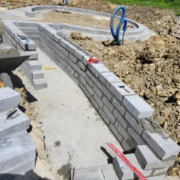 Wznoszenie muru z szarych bloczków betonowych na fundamencie, widoczne narzędzia murarskie, poziomica, taczka i fragment wykopu pod basen lub oczko wodne w tle.