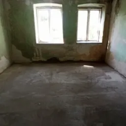 Wnętrze pustego pokoju z dwoma oknami, ściany z widocznymi śladami farby i uszkodzeniami, surowy beton na podłodze, widoczne pozostałości po instalacji na suficie.