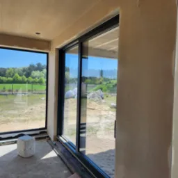 Świeżo nałożony, surowy tynk na ścianach wewnątrz budynku w Turku, widoczne duże okno z czarną ramą i przesuwne drzwi balkonowe, na podłodze zabezpieczenie.