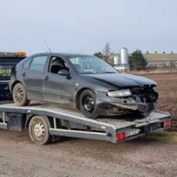Uszkodzony, czarny samochód osobowy na platformie lawety z włączonym pomarańczowym światłem ostrzegawczym, na tle wiejskiego krajobrazu z budynkami gospodarczymi i polem.