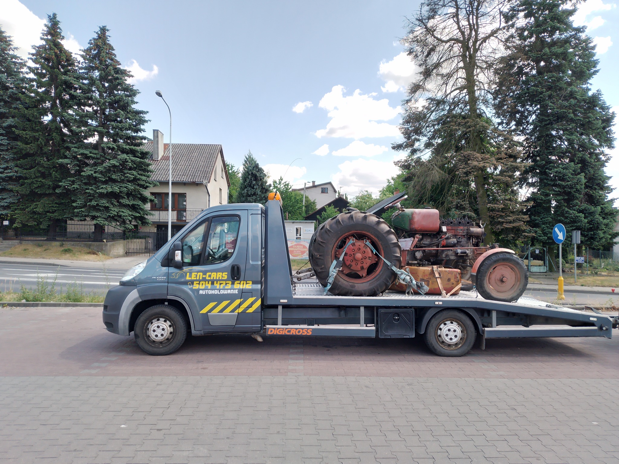 Szara laweta z pomarańczowym traktorem zabezpieczonym pasami transportowymi, widok z boku na tle ulicy i budynków.