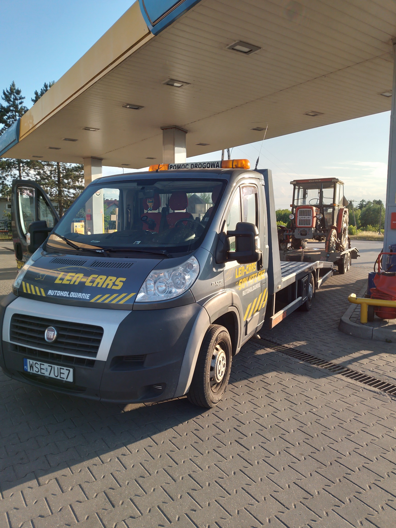 Szara laweta marki Fiat z żółtymi pasami i napisem 'LEN-CARS AUTOHOLOWANIE' na masce, przewożąca czerwony traktor Ursus na przyczepie, stojąca pod zadaszeniem stacji benzynowej. Otwarta kabina...