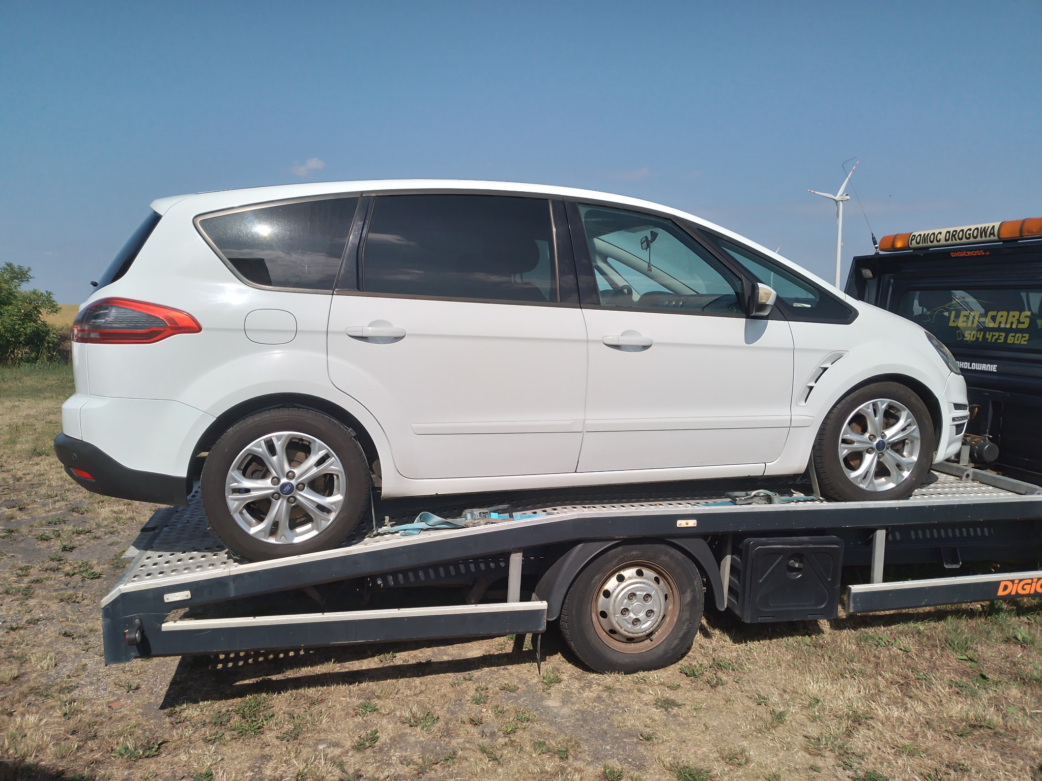 Biały samochód osobowy Ford S-Max zabezpieczony pasami na platformie lawety, w tle widoczny wiatrak turbiny wiatrowej.