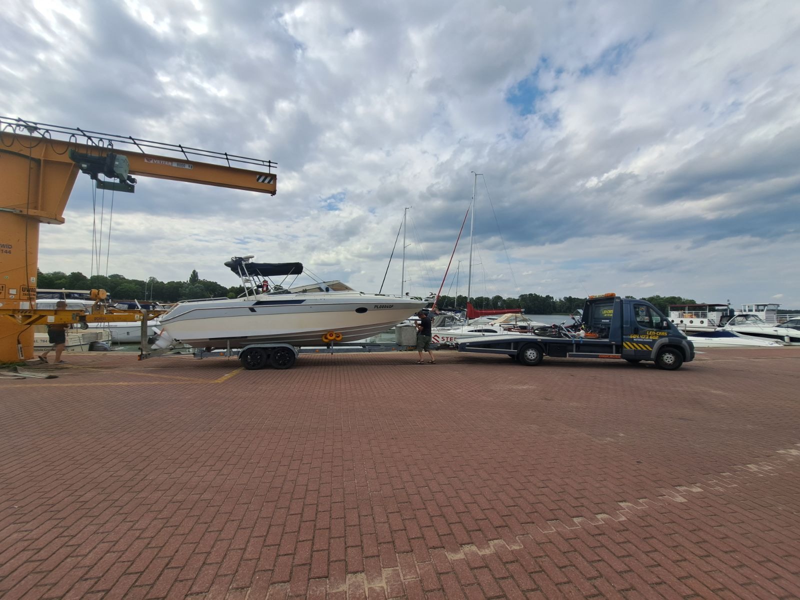 Biała motorówka z czarnym daszkiem, o nazwie 'PLOB040F', transportowana na lawecie przez niebieską ciężarówkę z dźwigiem w tle, na czerwonym brukowanym placu.