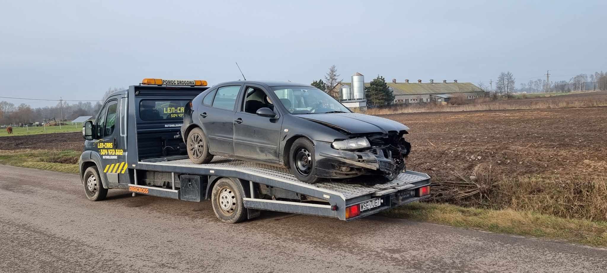 Uszkodzony, czarny samochód osobowy na platformie lawety z włączonym pomarańczowym światłem ostrzegawczym, na tle wiejskiego krajobrazu z budynkami gospodarczymi i polem.