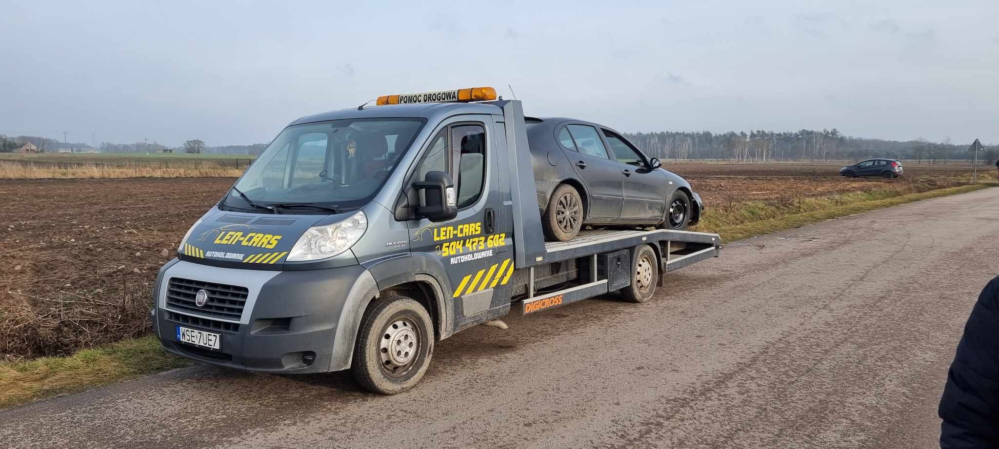 Szara laweta z napisem 'LEN-CARS' i numerem telefonu, przewożąca uszkodzony, szary samochód osobowy na tle pola i drzew, w pochmurny dzień.