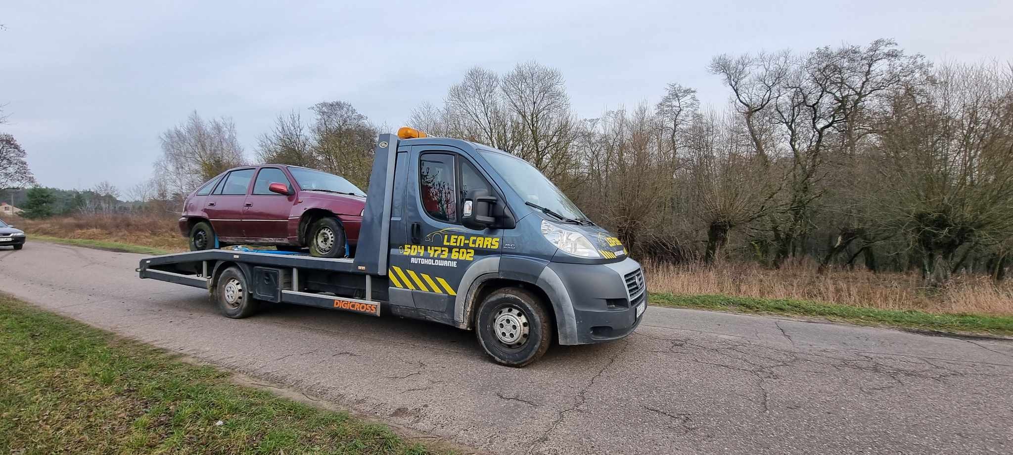 Szara laweta z czerwonym samochodem osobowym na platformie, zaparkowana na poboczu drogi wiejskiej w pochmurny dzień. Na kabinie widoczne logo firmy autoholowniczej i numer telefonu.