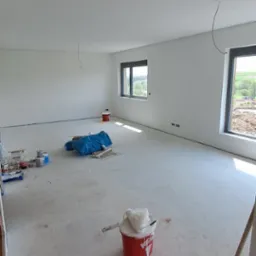 Świeżo pomalowane, puste pomieszczenie z dwoma oknami, widoczne ślady farby na podłodze, wiadro z farbą KREISEL, pędzel, szczotka i zabezpieczenia podłogi folią.