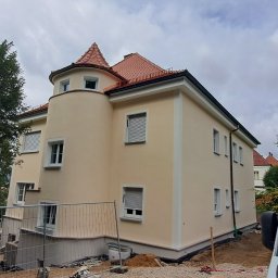 Usługi budowlane Łukasz Wójtowicz - Jasna elewacja dwupiętrowego domu z wieżyczką, widoczna z poziomu gruntu, z fragmentem ogrodzenia budowlanego i samochodu dostawczego w kadrze.