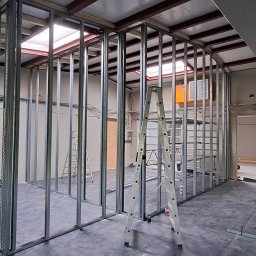 Usługi budowlane Łukasz Wójtowicz - Wnętrze w trakcie budowy ścian działowych z metalowych profili, widoczna drabina, surowy stan wykończenia, oświetlenie górne, podłoga zabezpieczona folią.