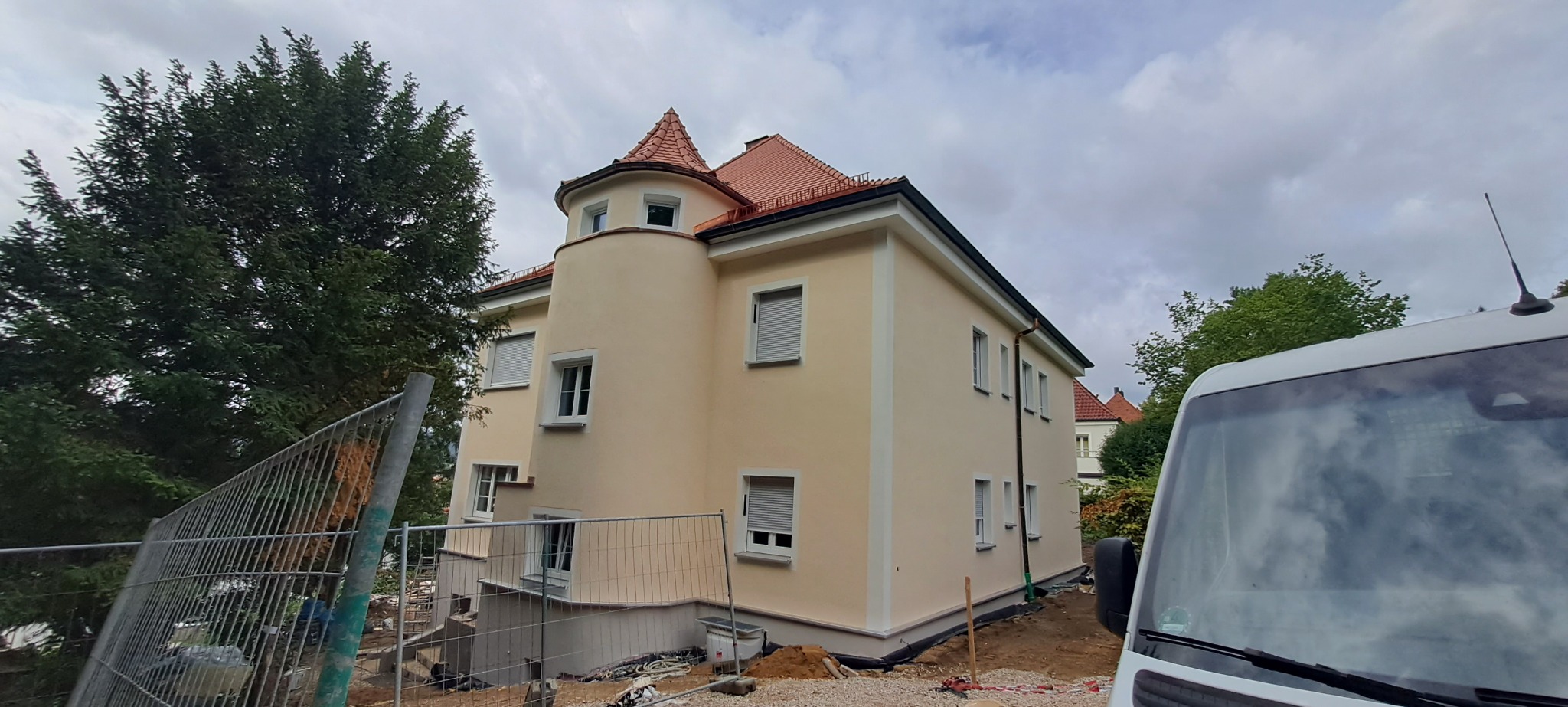 Jasna elewacja dwupiętrowego domu z wieżyczką, widoczna z poziomu gruntu, z fragmentem ogrodzenia budowlanego i samochodu dostawczego w kadrze.