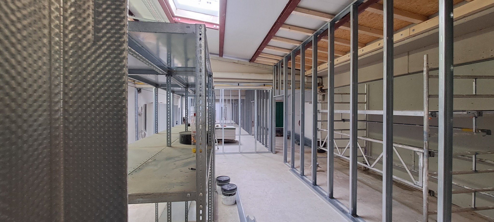 Wnętrze w trakcie prac wykończeniowych: stalowe profile konstrukcyjne ścian działowych na podłodze, widoczne instalacje, metalowe regały magazynowe z farbami w tle, oświetlenie sufitowe.