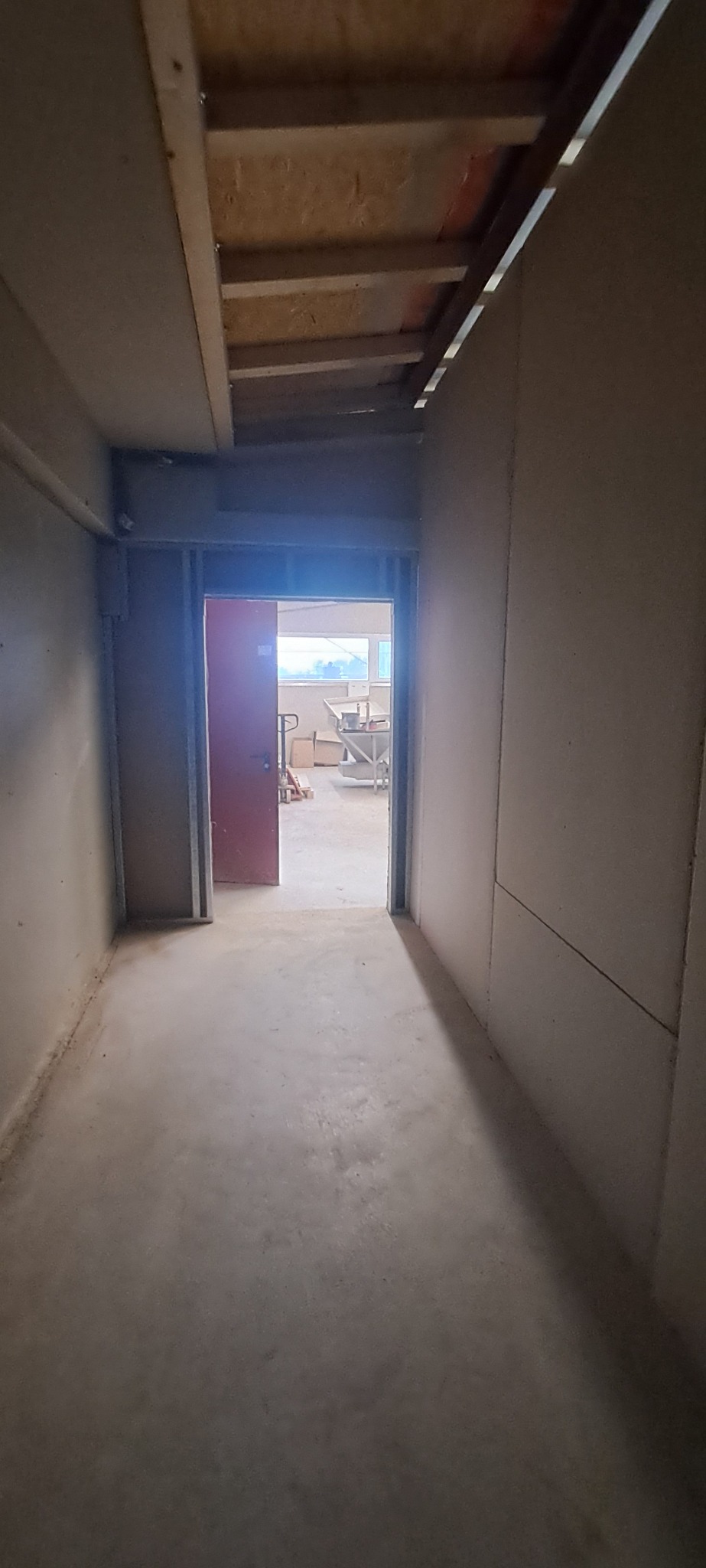 Wnętrze w trakcie prac wykończeniowych: surowy beton na podłodze, ściany obłożone płytami gipsowo-kartonowymi, widoczne elementy konstrukcji sufitu i otwarte przejście do kolejnego pomieszczenia...