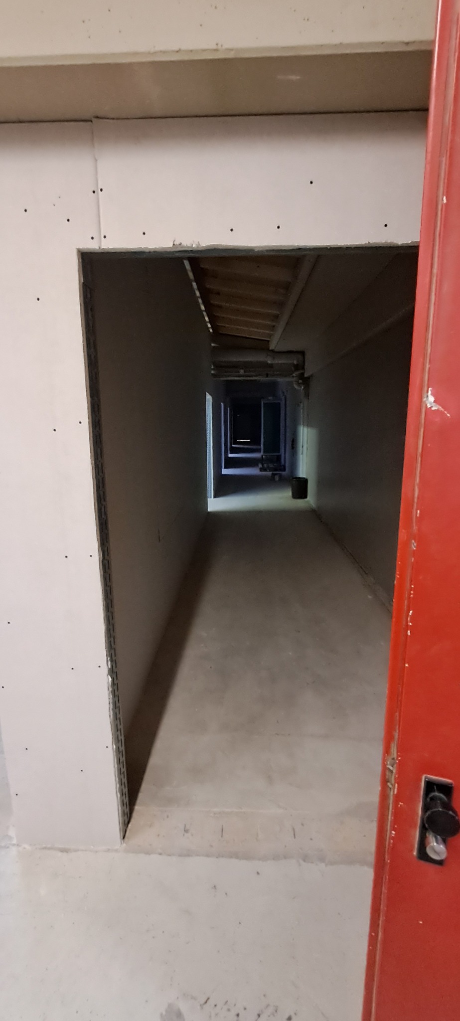 Korytarz w trakcie remontu z zamontowaną płytą kartonowo-gipsową na ścianach, widoczne niedoskonałości i łączenia płyt, czerwone drzwi po prawej stronie kadru.