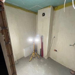 Mateusz Jadczak - Wnętrze w trakcie remontu: surowy stan z sufitami z płyt kartonowo-gipsowych, ściany przygotowane do malowania, widoczne instalacje elektryczne, halogen na statywie oświetla przestrzeń.