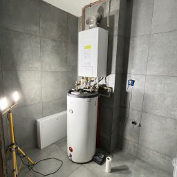 Mateusz Jadczak - Montaż kotła gazowego i zasobnika ciepłej wody w łazience wykończonej szarymi płytkami, widoczne elementy instalacji miedzianej i narzędzia pracy.