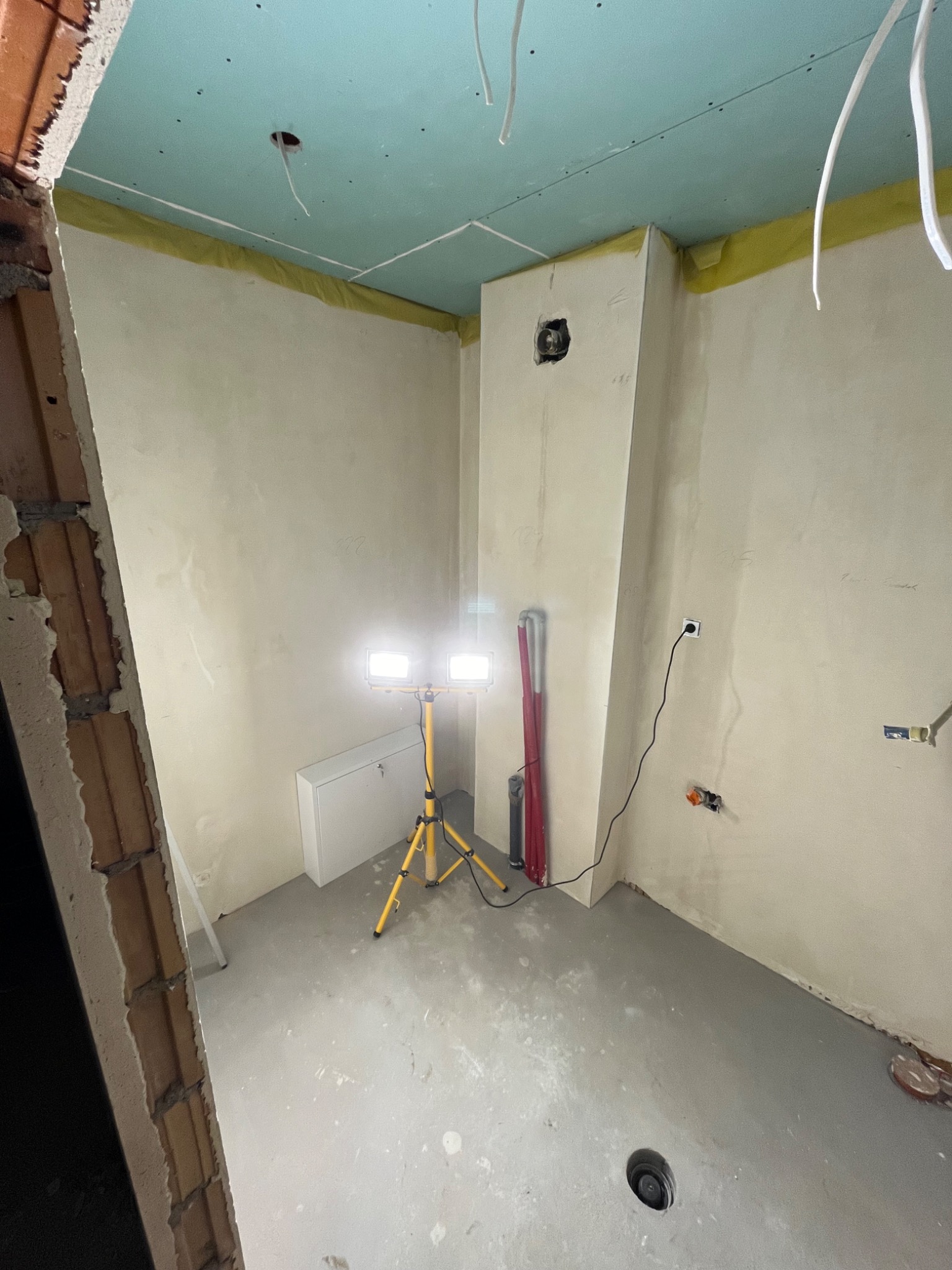 Wnętrze w trakcie remontu: surowy stan z sufitami z płyt kartonowo-gipsowych, ściany przygotowane do malowania, widoczne instalacje elektryczne, halogen na statywie oświetla przestrzeń.