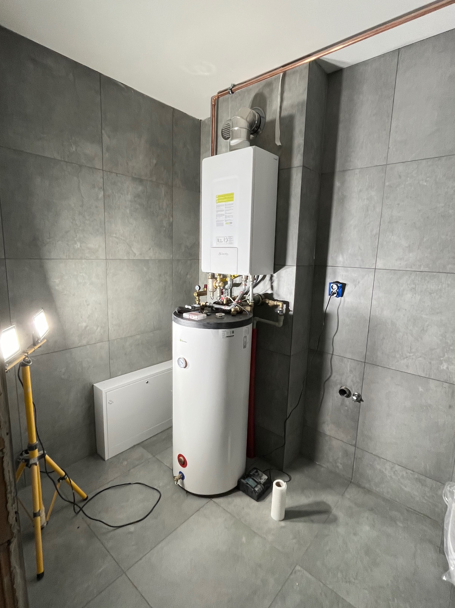 Montaż kotła gazowego i zasobnika ciepłej wody w łazience wykończonej szarymi płytkami, widoczne elementy instalacji miedzianej i narzędzia pracy.