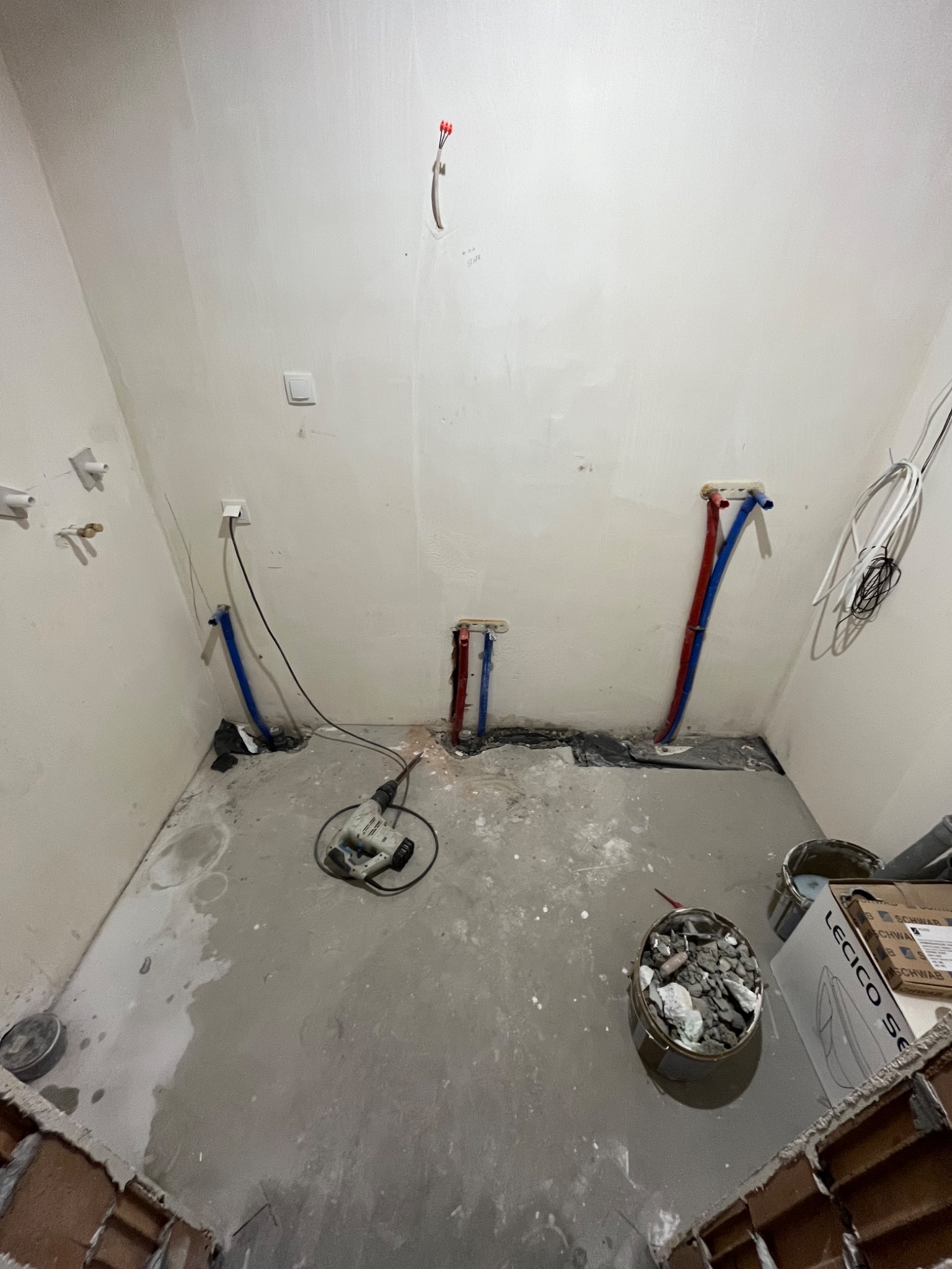 Wnętrze łazienki w trakcie prac remontowych; widoczne wyprowadzone instalacje wodne (rury czerwone i niebieskie), kable elektryczne, ślady po kuciu w ścianach, oraz narzędzia pracy (wiertarka...