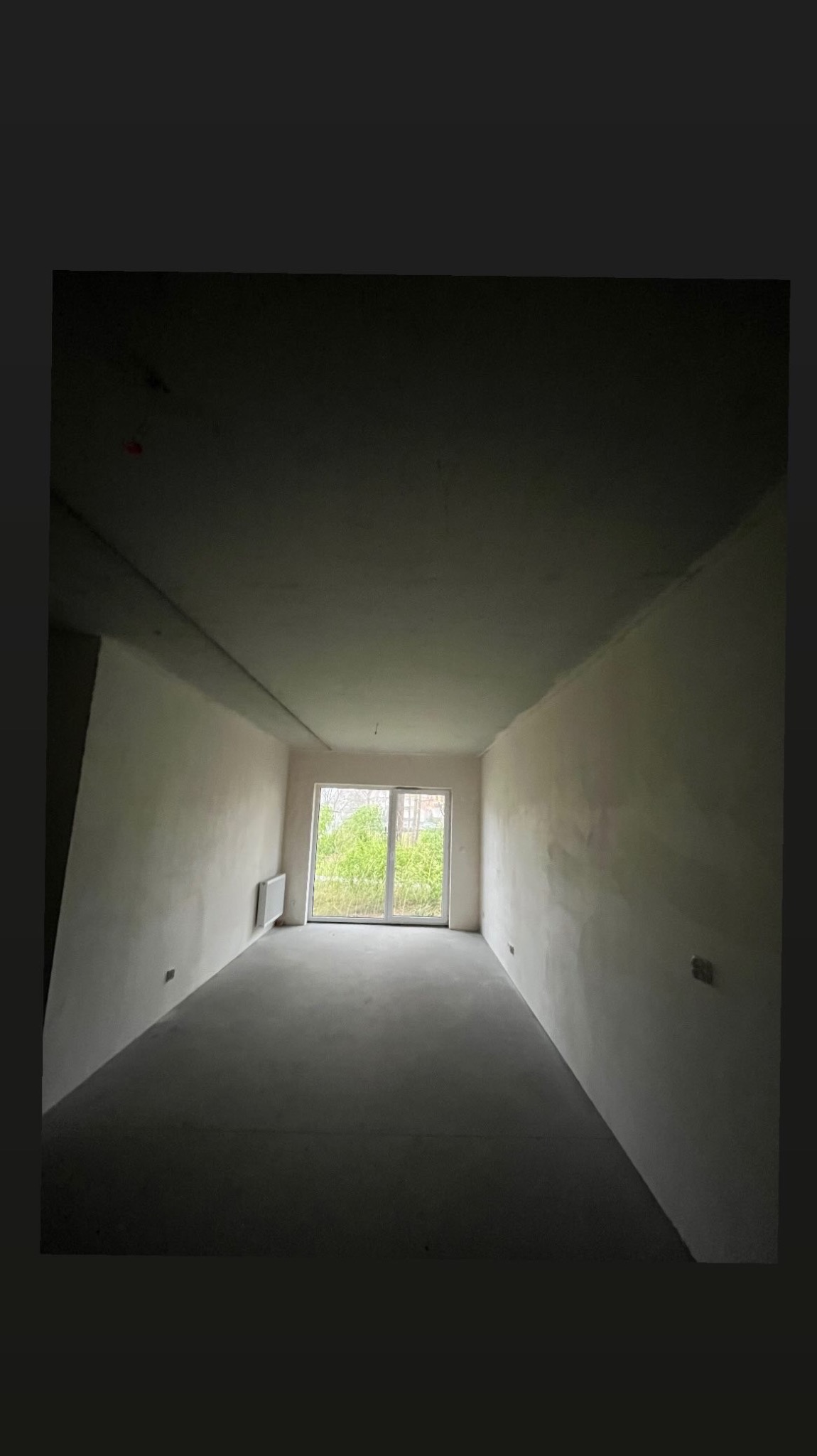 Surowe wnętrze pokoju w stanie deweloperskim z widocznymi śladami po gładzi na ścianach, betonową podłogą i grzejnikiem pod oknem balkonowym.