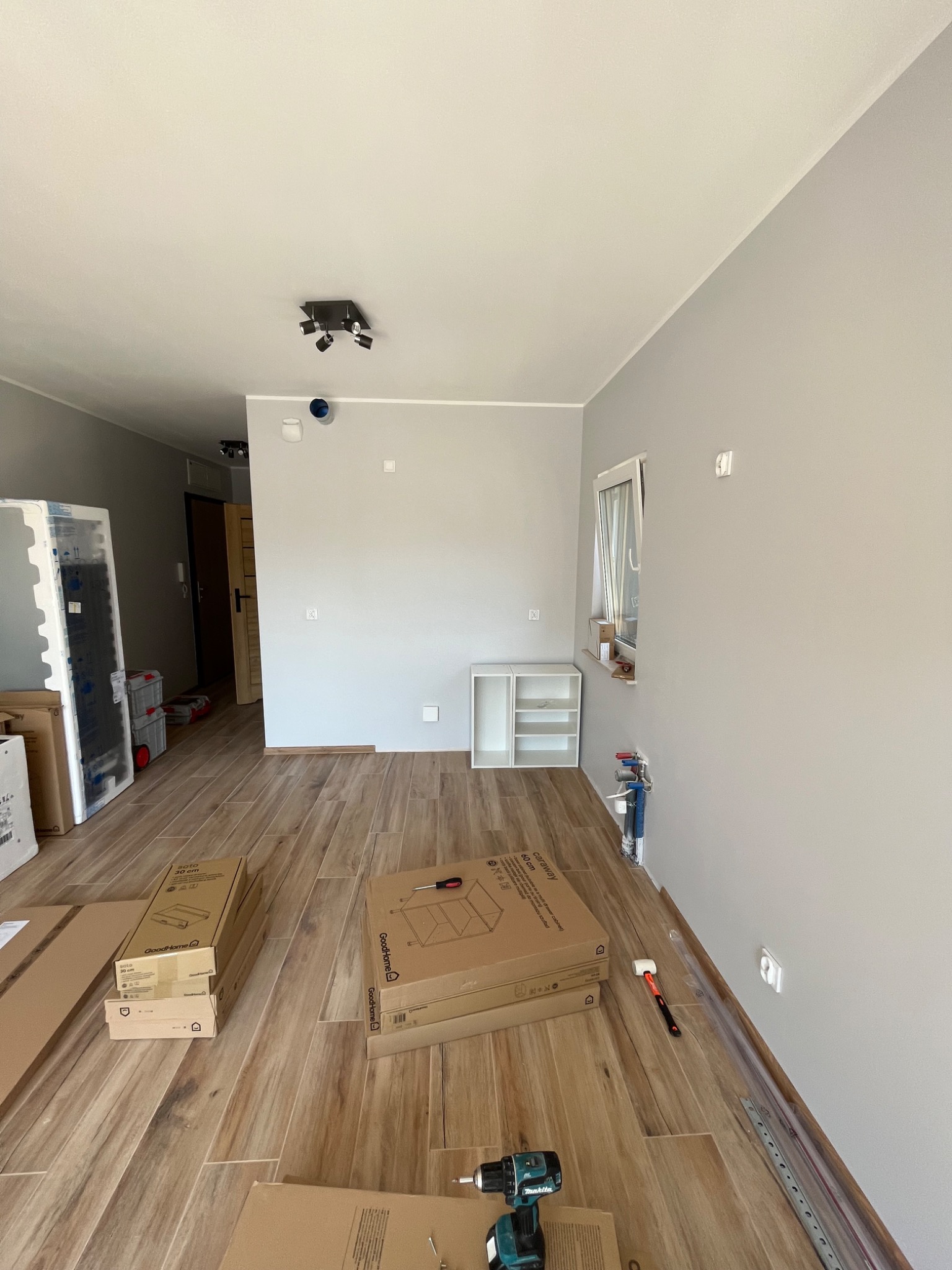 Pokój w trakcie wykańczania: drewnopodobne płytki na podłodze, szare ściany, częściowo złożone białe szafki, kartony meblowe, wiertarka i młotek gumowy widoczne na podłodze.