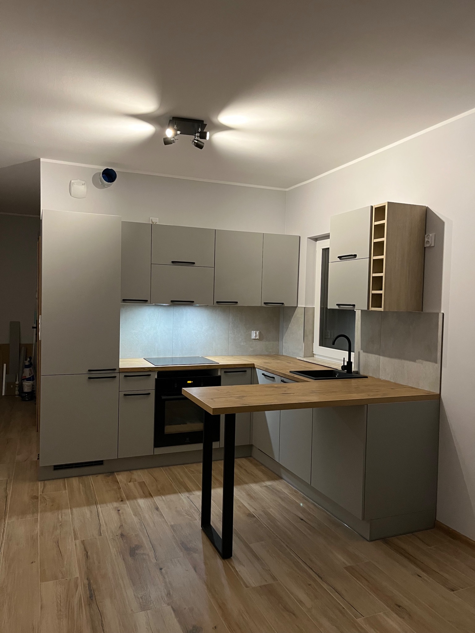 Nowoczesna kuchnia w szarościach z drewnianymi akcentami, oświetleniem punktowym i czarną armaturą, widok na zabudowę kuchenną z blatem i barkiem.