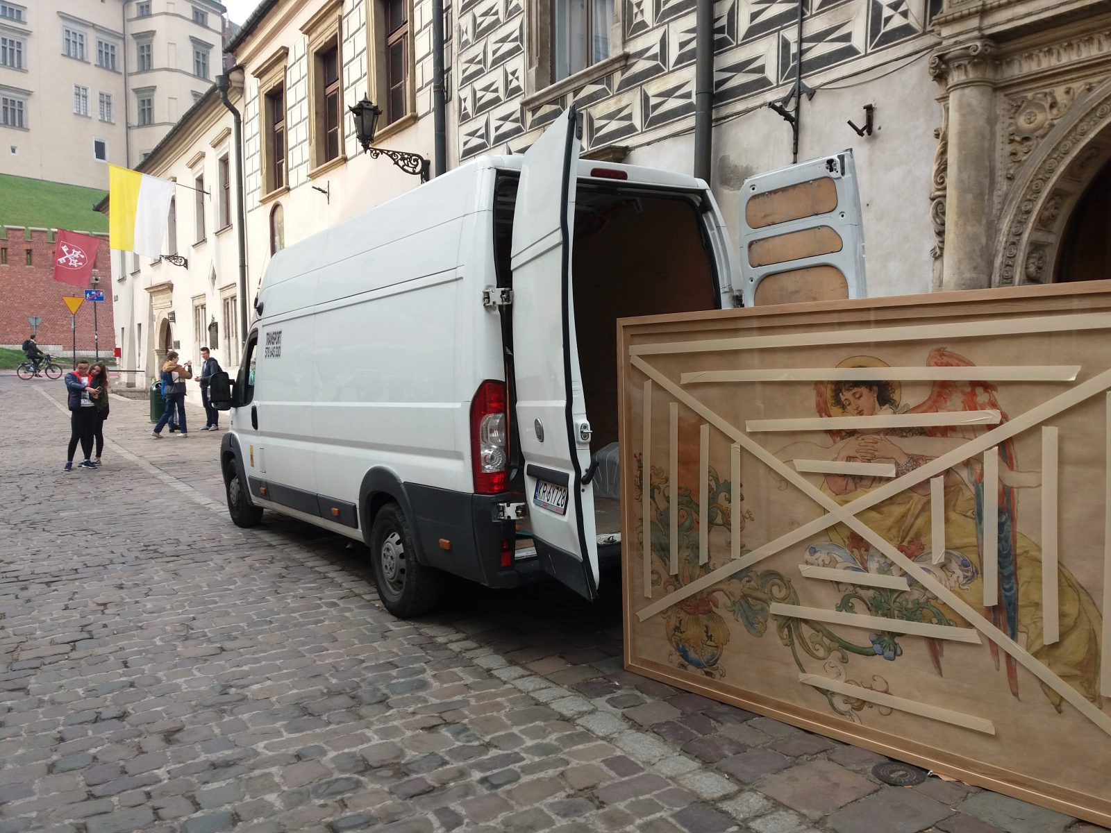 Biały van z otwartymi drzwiami bagażnika na tle brukowanej ulicy i zabytkowej architektury; w bagażniku widoczny obraz zabezpieczony taśmą malarską, gotowy do transportu.