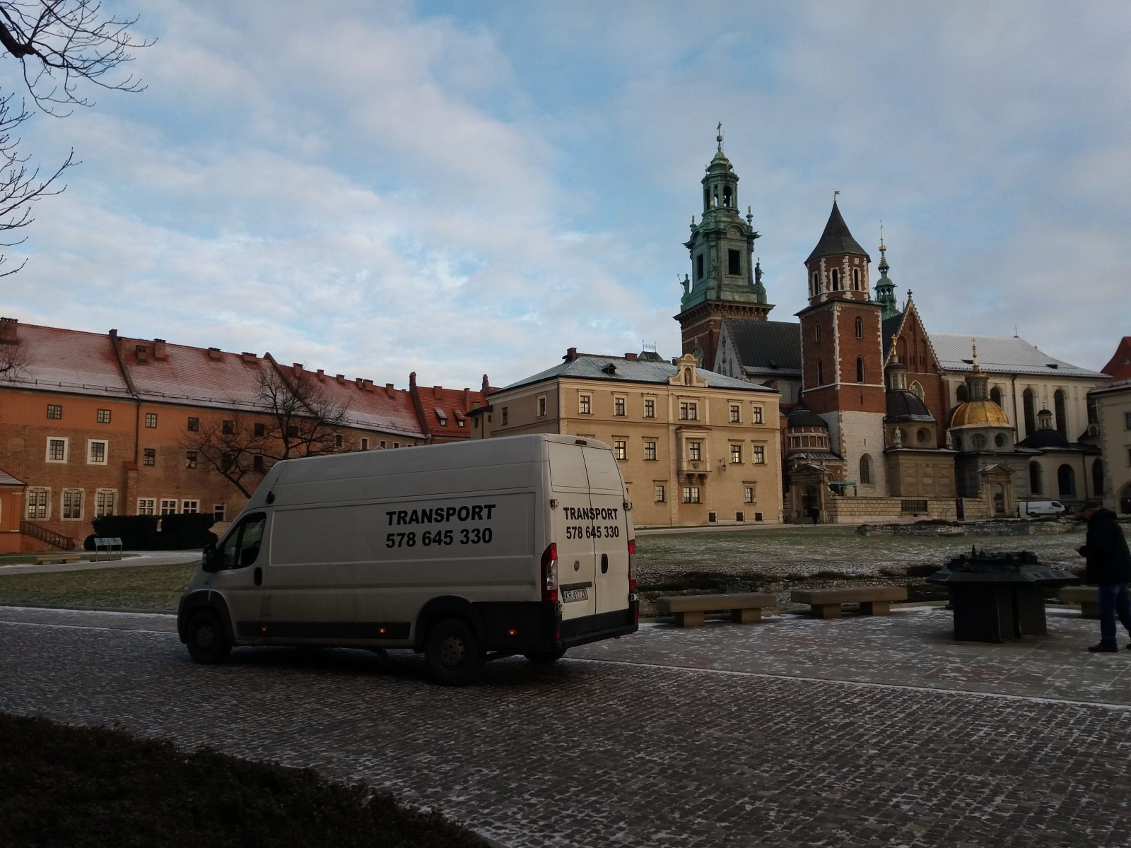 Biały samochód dostawczy z napisem 'TRANSPORT 578 645 330' zaparkowany na tle Wawelu w Krakowie.