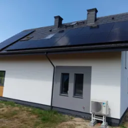 Nowoczesny dom jednorodzinny z zainstalowanymi panelami fotowoltaicznymi na dachu z ciemnej dachówki, klimatyzator Daikin na elewacji, widok z boku.