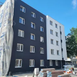 Wielopiętrowy budynek z elewacją w kolorze grafitowym i białym, część budynku w trakcie prac, osłonięta plandeką z logo firmy, widoczne rusztowania i materiały budowlane.