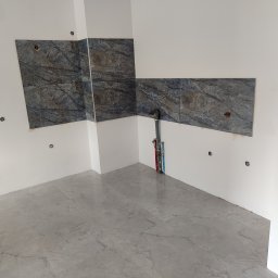 BurbanBud - Fragment łazienki w trakcie prac wykończeniowych: położone szare płytki podłogowe z wzorem marmuru, ściany częściowo wyłożone ciemnymi płytkami imitującymi kamień, widoczne rury i instalacje...