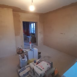 BurbanBud - Pokój w trakcie prac wykończeniowych: ściany po gładzi, worki z klejem na podłodze, widoczne narzędzia i materiały budowlane. W tle widoczne okno i inne pomieszczenie.