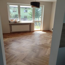 BurbanBud - Jasna, drewniana podłoga ułożona w jodełkę w pustym pokoju z oknem i grzejnikiem. Widok na zewnątrz przez okno, lampa na suficie.