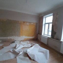 BurbanBud - Pokój w trakcie przygotowań do remontu: zerwane tapety, odsłonięte fragmenty ścian, widoczne ślady po farbie i kleju. Okno z grzejnikiem w tle. Bałagan na podłodze.