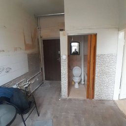 BurbanBud - Wnętrze mieszkania w trakcie remontu: widoczne ściany przygotowane do malowania, fragmenty starej glazury, drzwi wejściowe i wejście do toalety z lustrem.